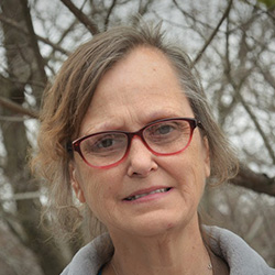 Image of Dee Banta. Dee is wearing pink glasses. Smiling at the camera. There are bare tree branches in the background.