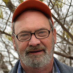 Image of Kevin Wilson. Kevin is wearing glasses and a orange baseball hat. Smiling to the camera. There are bare tree branches in the background.