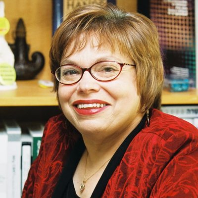 Judith Heumann has short brown hair and oval glasses. She is nicely dressed in a red top.